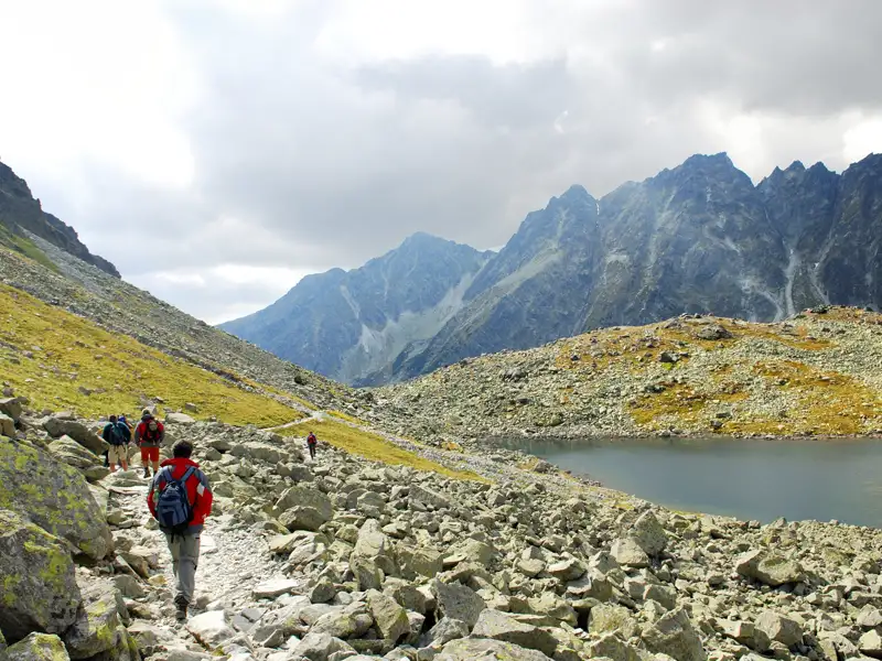 Wanderer auf einem steinigen Weg in den Bergen, mit einem Bergsee und Gipfeln im Hintergrund.