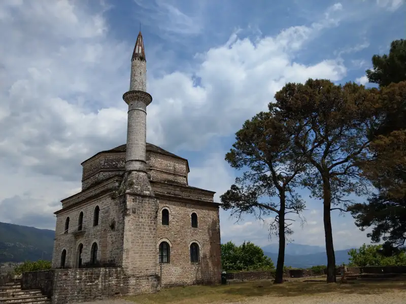 Alte Steinmoschee mit Minarett, umgeben von Bäumen und mit Bergen im Hintergrund.