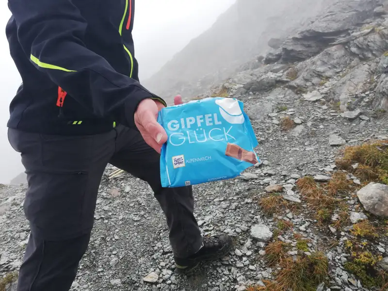Wanderer mit einer Packung "Gipfelglück" Alpenmilch Schokolade auf einem Bergpfad.