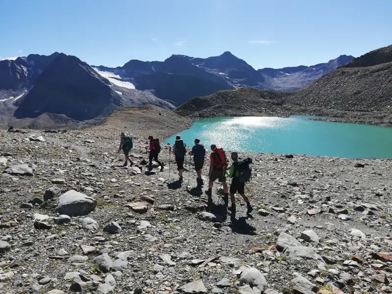 Wandergruppe auf einem Bergpfad neben einem türkisfarbenen See mit Bergen und Gletscher im Hintergrund.