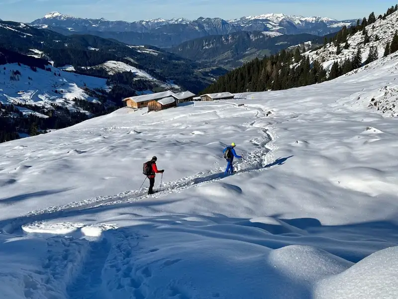 Zwei Schneeschuhwanderer auf einem verschneiten Bergpfad. Eine Hütte ist in der Ferne sichtbar, ebenso wie die umliegenden Berge.