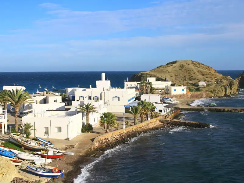 Blick auf einen Küstenort mit weißen Häusern und Fischerbooten am Meer. Im Hintergrund ist eine kleine Insel zu sehen.