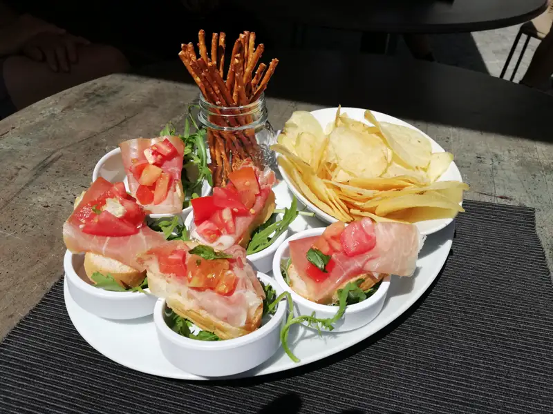 Bruschetta mit Tomaten, Schinken und Rucola, serviert mit Kartoffelchips und Salzstangen.