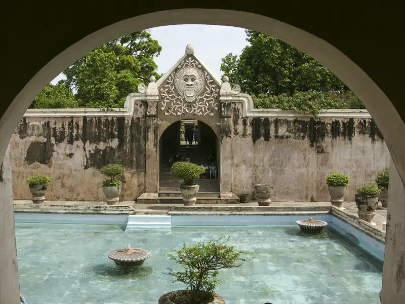 Ein Wassergarten mit Springbrunnen und einem kunstvollen Tor, gesehen durch einen Torbogen.