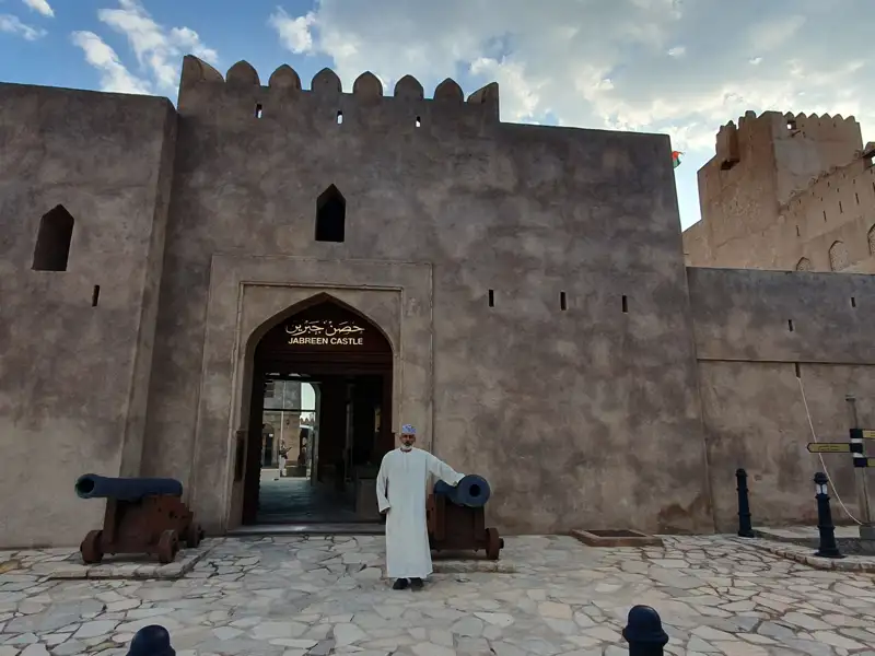 Jabreen Castle Eingang mit Kanonen und einer Person in traditioneller Kleidung.