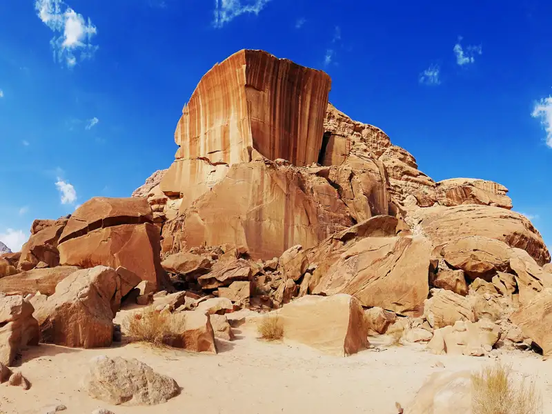 Eine große, rotbraune Felsformation ragt in den blauen Himmel. Am Fuß der Formation liegen weitere Felsen und Sand.