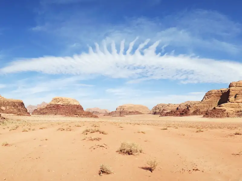 Weitläufige Wüstenlandschaft mit roten Felsformationen und einer einzigartigen Wolkenformation am blauen Himmel.