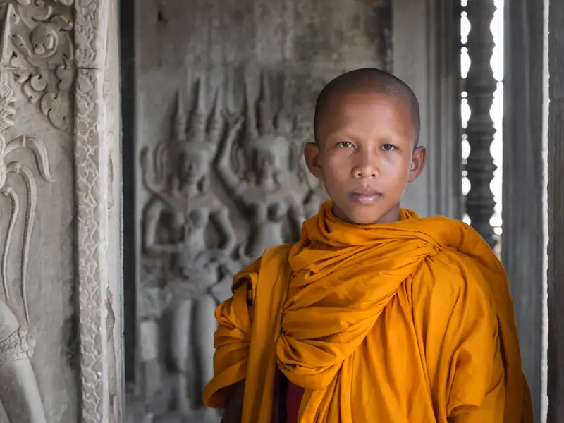 Junger Mönch in traditioneller orangefarbener Robe vor kunstvollen Steinreliefs in einem Tempel.