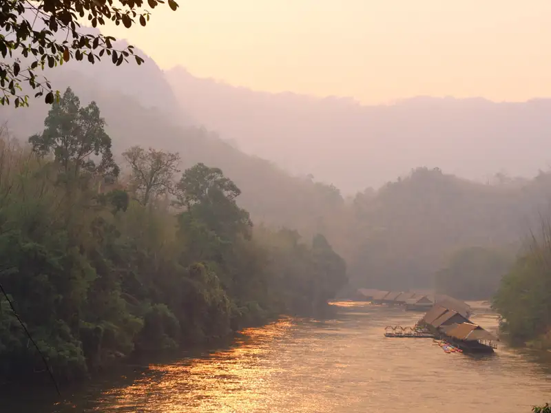Schwimmende Unterkünfte auf einem Fluss bei Sonnenuntergang, umgeben von üppiger Vegetation und sanften Hügeln.