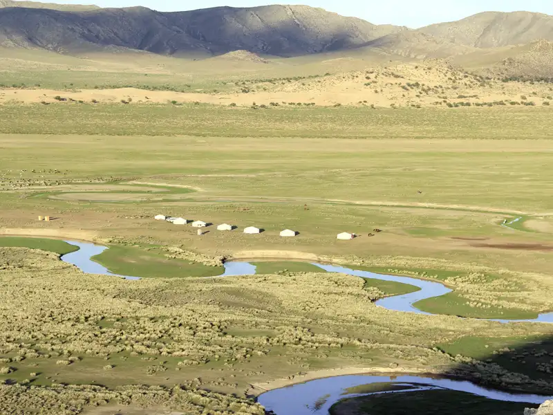 Traditionelles Jurtencamp in einer mongolischen Landschaft mit Fluss und Bergen im Hintergrund.