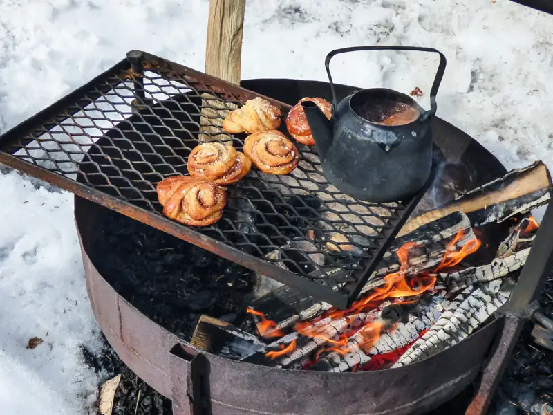 Zubereitung von Zimtschnecken und einem Heißgetränk über einem offenen Feuer im Schnee.