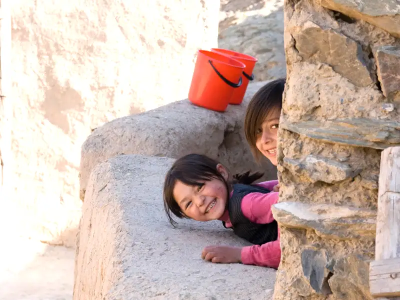 Zwei Kinder, die sich hinter einer Mauer verstecken, lächeln in die Kamera. Zwei rote Eimer stehen auf der Mauer.