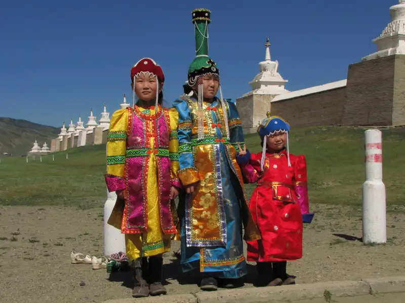 Drei Kinder in traditioneller mongolischer Kleidung vor einer Reihe von weißen Stupas in der Mongolei.