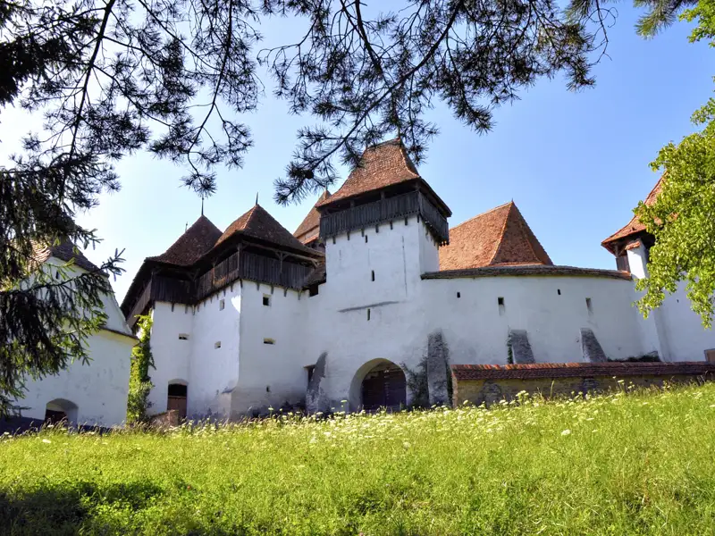 Historische Wehrkirche mit mehreren Türmen und weißer Mauer, umgeben von einer grünen Wiese.