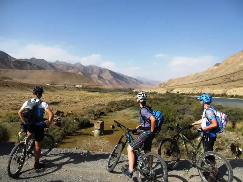 Drei Mountainbiker mit Rucksäcken und Helmen halten an einem Aussichtspunkt an und betrachten die Landschaft mit Fluss und Bergen.
