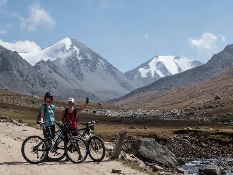 Zwei Personen auf Mountainbikes pausieren auf einem Bergpfad mit schneebedeckten Bergen im Hintergrund.