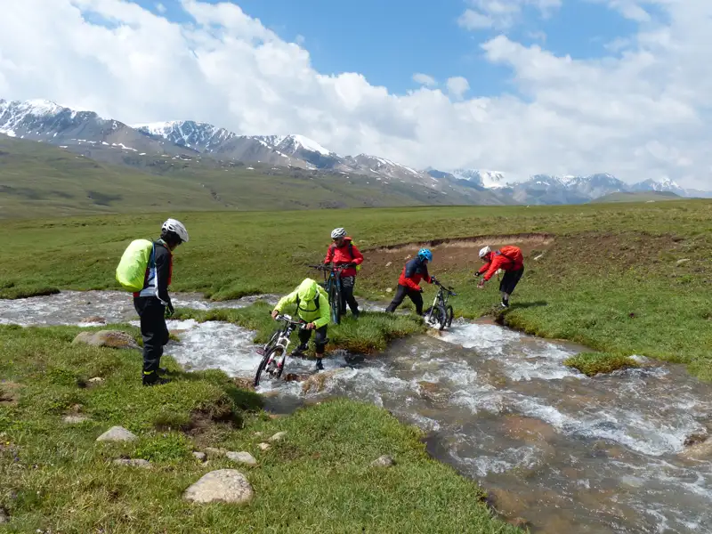 Mountainbiker schieben ihre Fahrräder durch einen Fluss in einer Gebirgslandschaft.