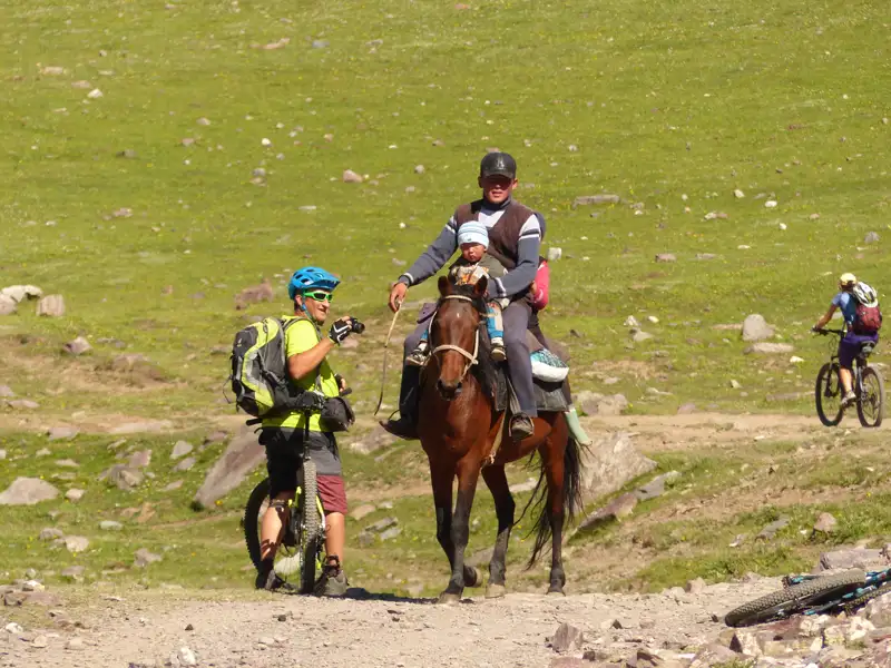 Reisende begegnen sich auf einem Weg: Ein Mann mit Kind auf dem Pferd und ein Radfahrer halten an, um miteinander zu sprechen. Weitere Radfahrer sind im Hintergrund zu sehen.