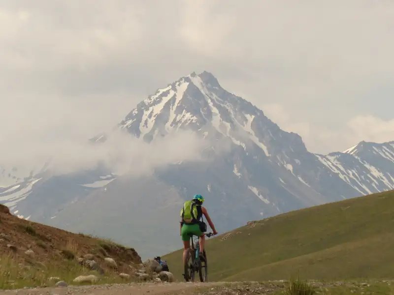 Mountainbiker auf einem Bergpfad mit schneebedecktem Gipfel im Hintergrund.