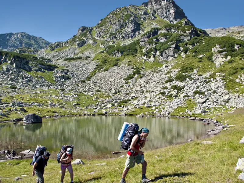 Drei Wanderer mit schweren Rucksäcken beim Trekking in den Bergen. Ein Bergsee und felsige Landschaft im Hintergrund.
