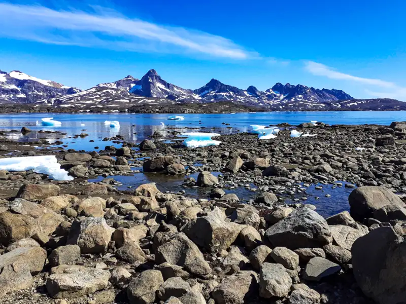 Felsiger Strand mit Eisbergen und schneebedeckten Bergen im Hintergrund.