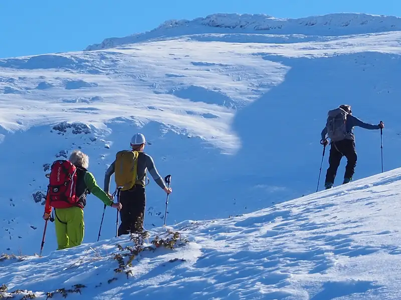 Drei Personen mit Skitouren-Ausrüstung steigen einen verschneiten Berg hinauf.
