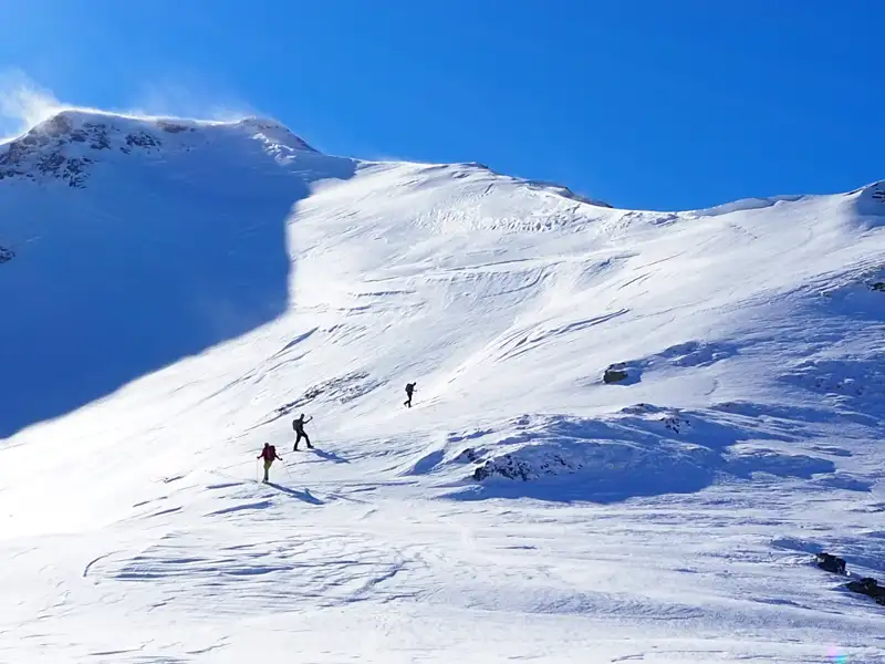Drei Personen auf Skitour in den Bergen. Der Schnee bedeckt den gesamten Hang, und der Gipfel ist im Hintergrund zu sehen.