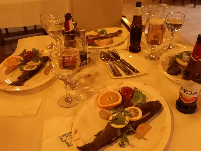 Gegrillter Fisch auf Tellern, angerichtet mit Zitronen, Orangen und Kräutern. Getränke wie Bier, Wasser und möglicherweise Wein begleiten das Essen.