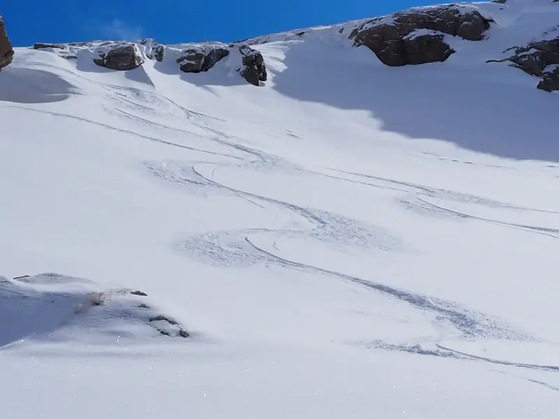 Abfahrtsspuren im frischen Schnee, umgeben von einer verschneiten Berglandschaft unter blauem Himmel.