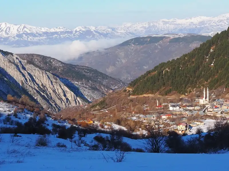 Blick auf ein verschneites Bergdorf im Tal, umgeben von schneebedeckten Bergen und Wäldern im Winter.