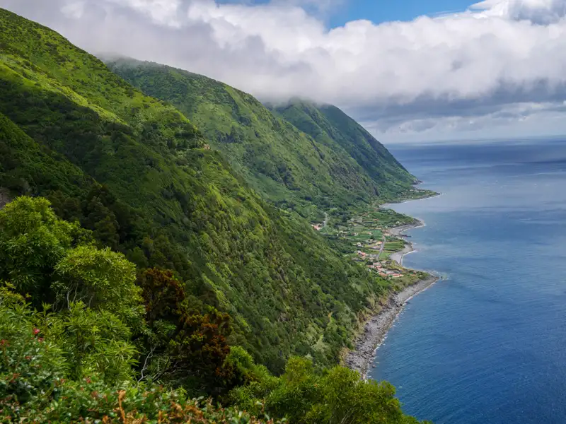 Steile, grüne Berghänge führen hinunter zum blauen Meer. Ein kleiner Küstenort ist sichtbar.