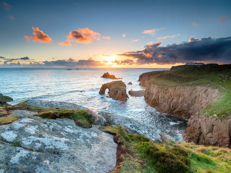 Sonnenuntergang an der felsigen Küste mit Blick auf einen natürlichen Felsbogen und einen Leuchtturm in der Ferne.
