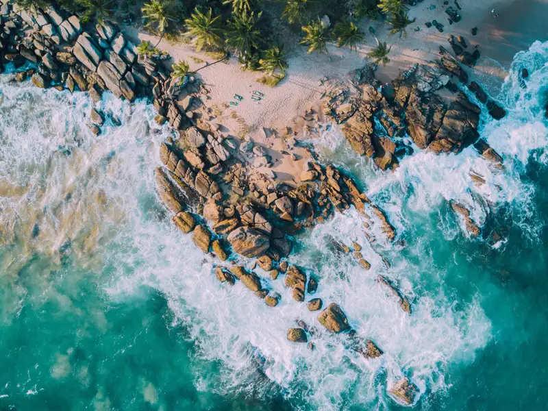 Luftaufnahme einer tropischen Küste: Palmengesäumter Sandstrand, Felsen und türkisfarbenes Wasser.