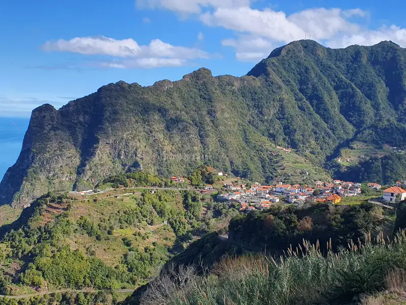 Panoramablick auf ein Dorf an der Küste, umgeben von grünen Bergen und dem Meer im Hintergrund.
