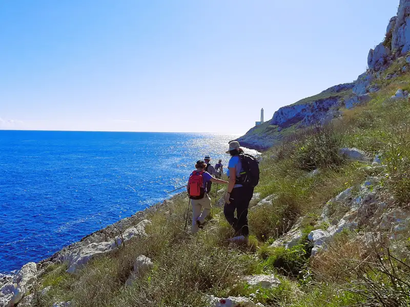 Wanderer auf einem Küstenpfad mit Blick auf das Meer und einen Leuchtturm in der Ferne.