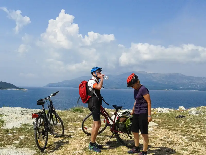 Zwei Radfahrer mit Helmen und Rucksäcken machen eine Pause an der Küste und trinken Wasser. Im Hintergrund ist das Meer und eine bergige Landschaft zu sehen.
