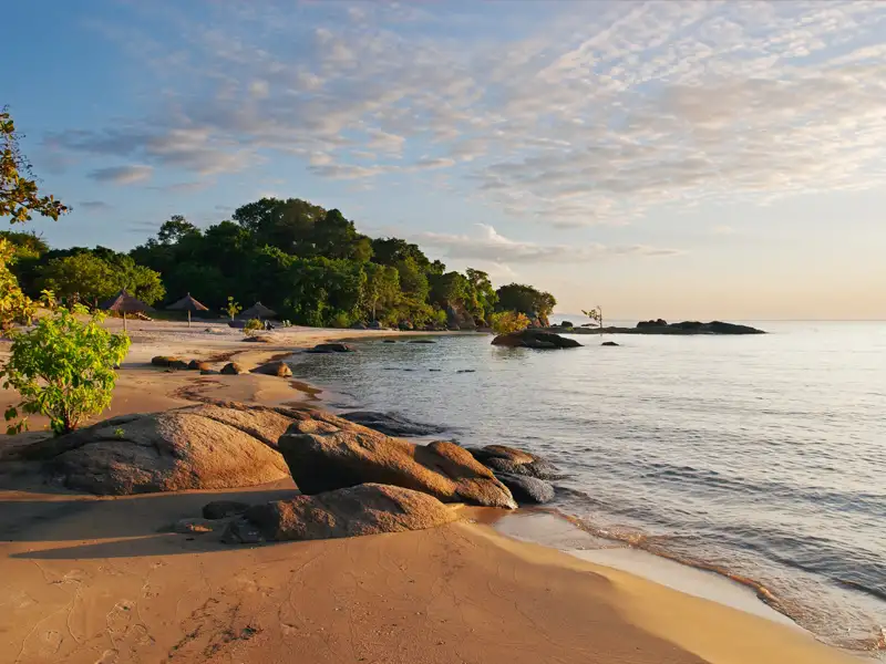 Ruhiger Strand mit goldenen Sand, Felsen und bewaldeter Küste.