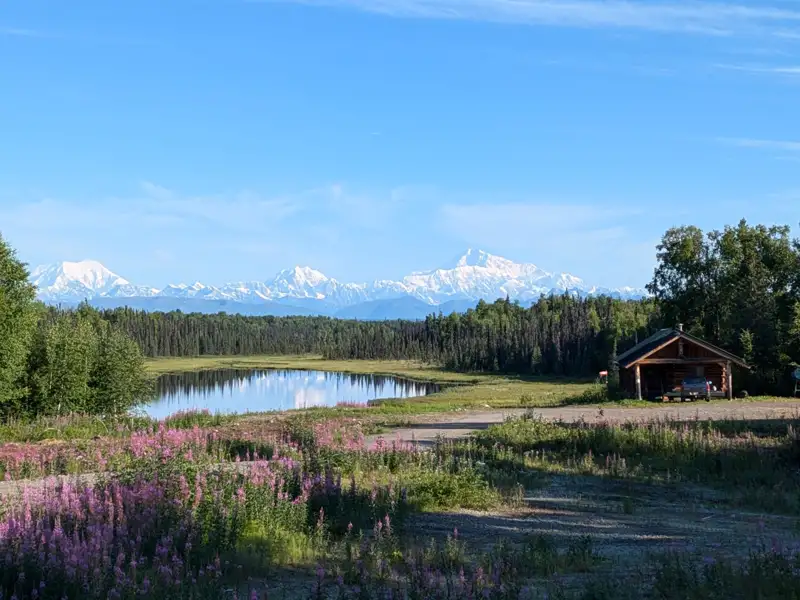 Idyllischer Bergsee mit einer Holzhütte und schneebedeckten Bergen im Hintergrund.
