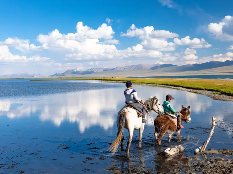 Zwei Kinder reiten auf einem Pferd und einem Esel durch einen See, umgeben von Bergen.