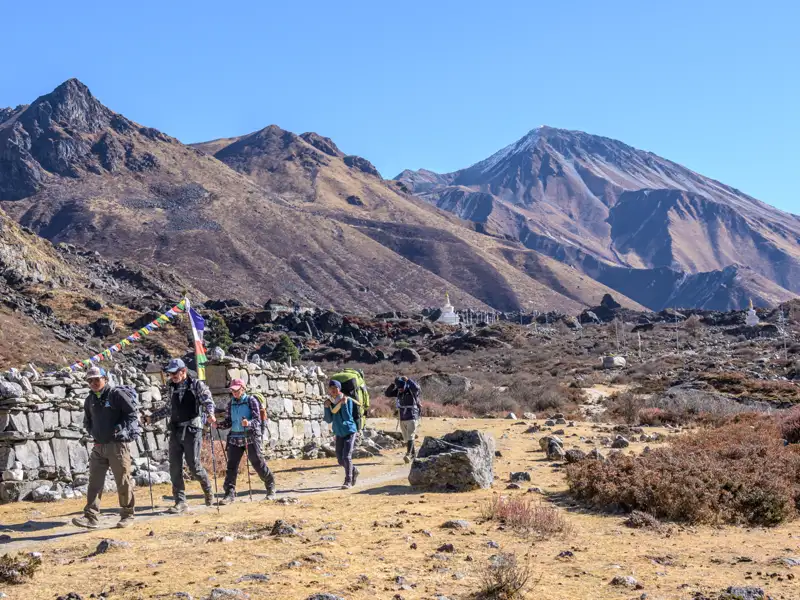 Wanderer auf einem Bergpfad mit Gebetsfahnen und Stupas im Hintergrund.