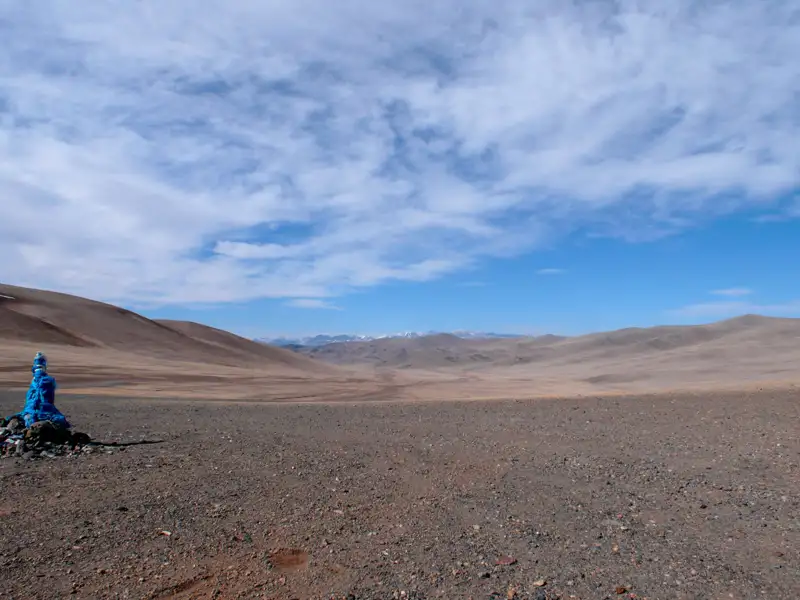 Blauer Gebetsfahnen-bedeckter Stein in einer trockenen Landschaft mit Hügeln und bewölktem Himmel.
