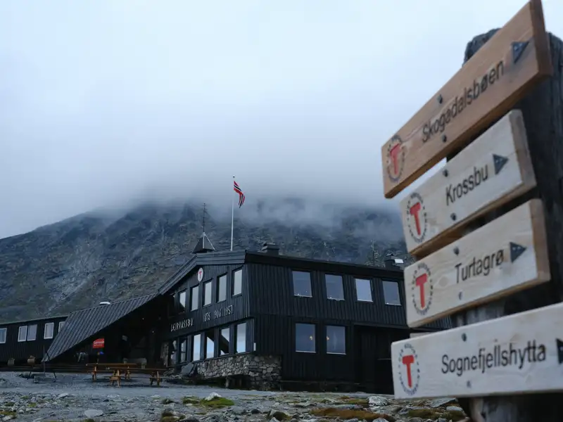 Die Sognefjellshytta, eine Berghütte in Norwegen, mit Wegweisern zu verschiedenen Wanderzielen. Nebel bedeckt die umliegenden Berge.