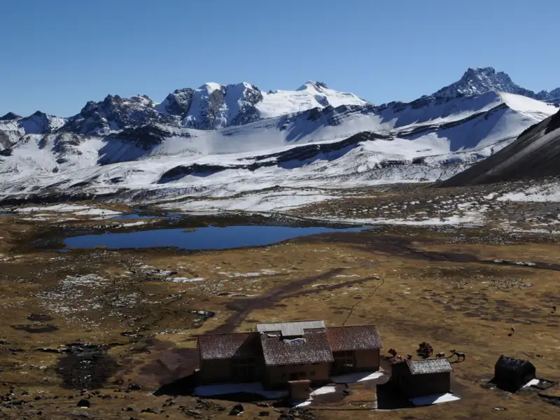Eine Berghütte bietet Unterkunft inmitten der Andenlandschaft mit einem See und schneebedeckten Gipfeln im Hintergrund.