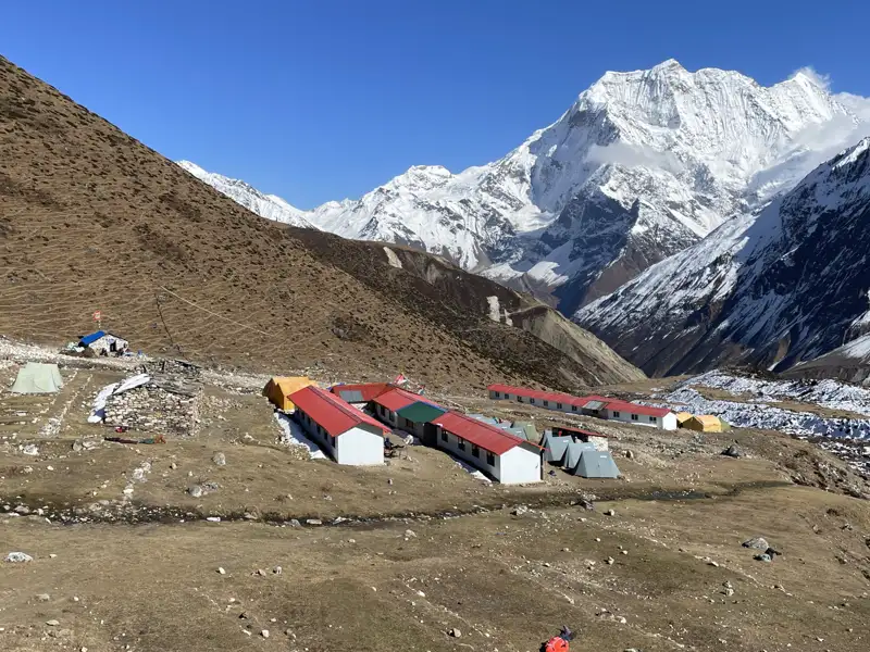 Basislager mit Unterkünften und Zelten inmitten einer Berglandschaft mit Blick auf einen schneebedeckten Gipfel.