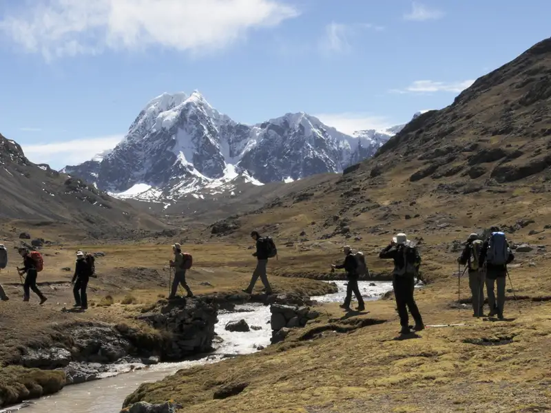 Wanderer überqueren einen Gebirgsbach in den Anden. Der schneebedeckte Gipfel im Hintergrund deutet auf die Höhe der Wanderung hin.