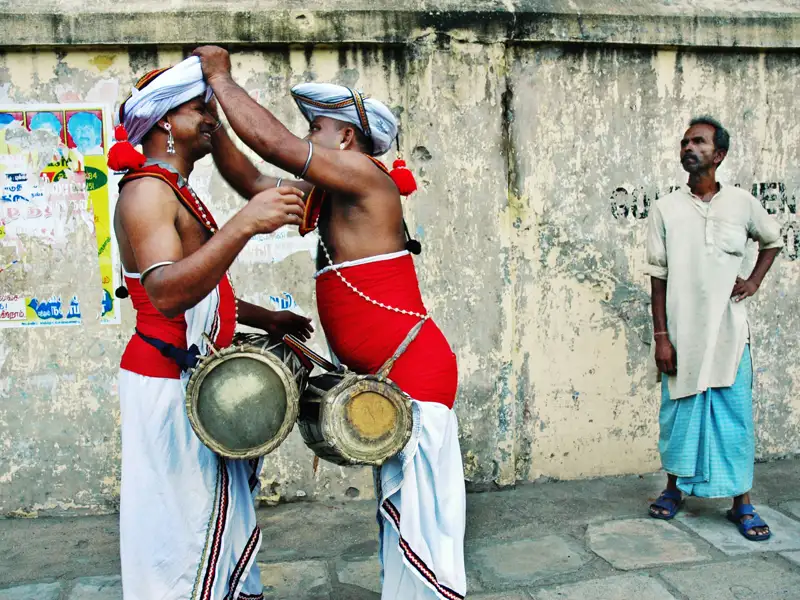 Zwei Musiker mit traditionellen Trommeln und roter und weißer Kleidung interagieren miteinander, während ein Zuschauer in blau-weißer Kleidung zusieht.