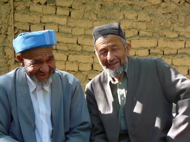 Zwei lächelnde Männer in traditioneller Kleidung vor einer Lehmziegelwand.