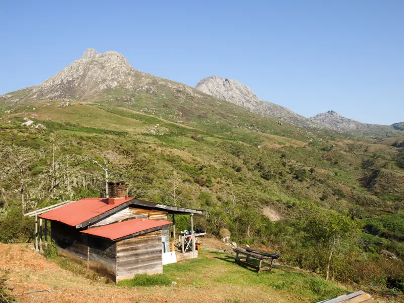 Eine einfache Berghütte mit rotem Dach inmitten einer grünen Hügellandschaft mit hohen Bergen im Hintergrund.