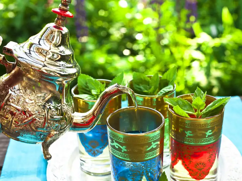 Zubereitung von marokkanischem Minztee: Eine silberne Teekanne giesst Tee in Gläser mit frischen Minzblättern.