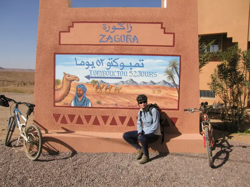 Radreise in Marokko: Pause in Zagora vor einem Schild mit der Aufschrift 'Timbuktu 52 Tage'.